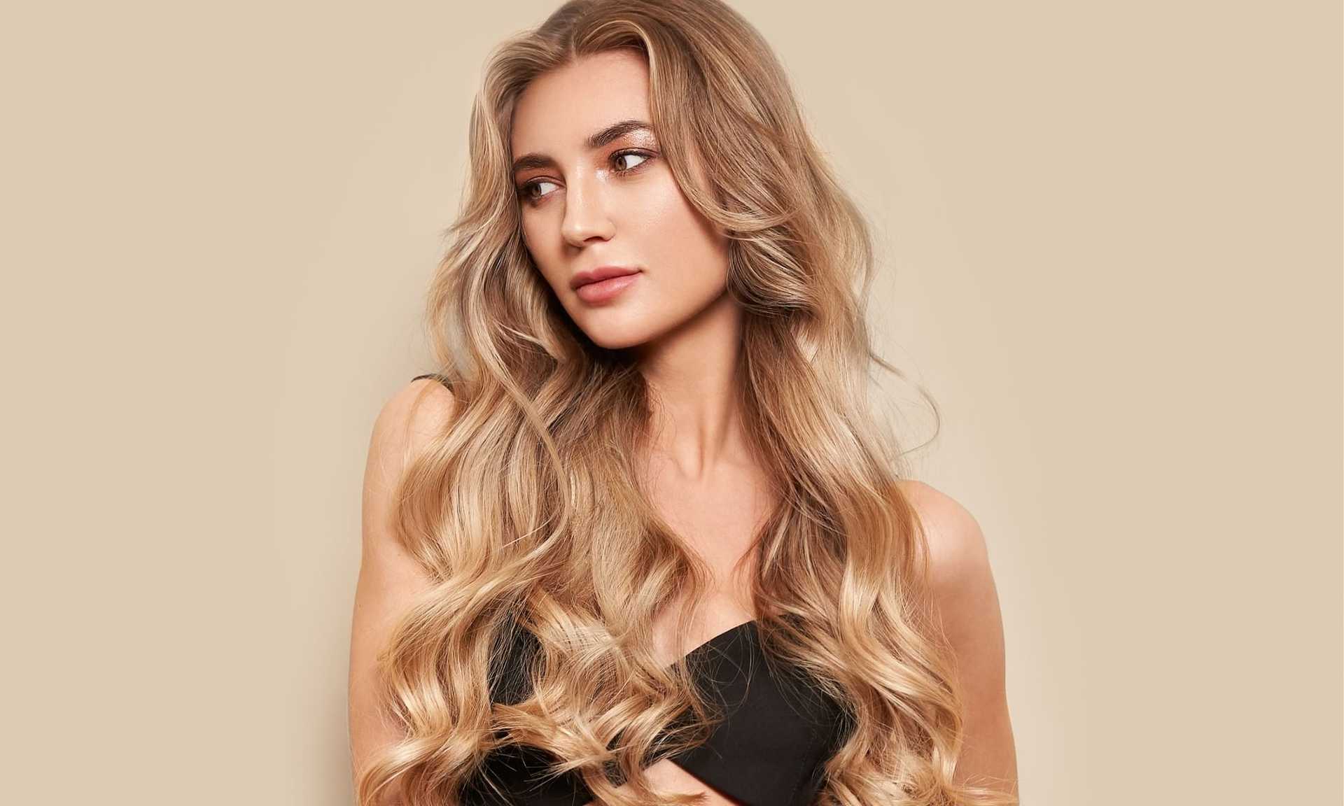 Woman with long wavy blonde hair, wearing a black top, looking to the side against a beige background.