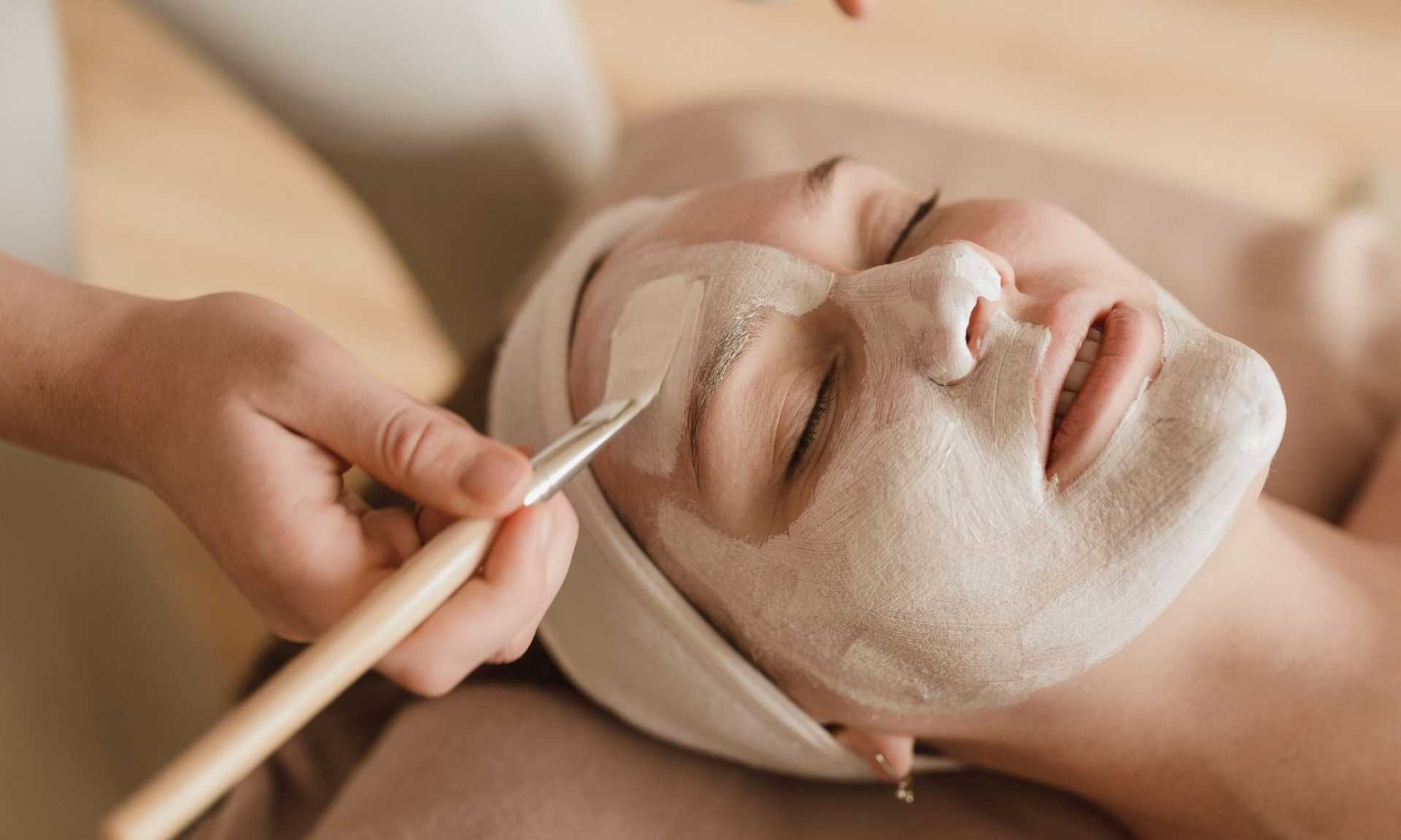 Person receiving a facial treatment with a brush applying a cream mask on their face.